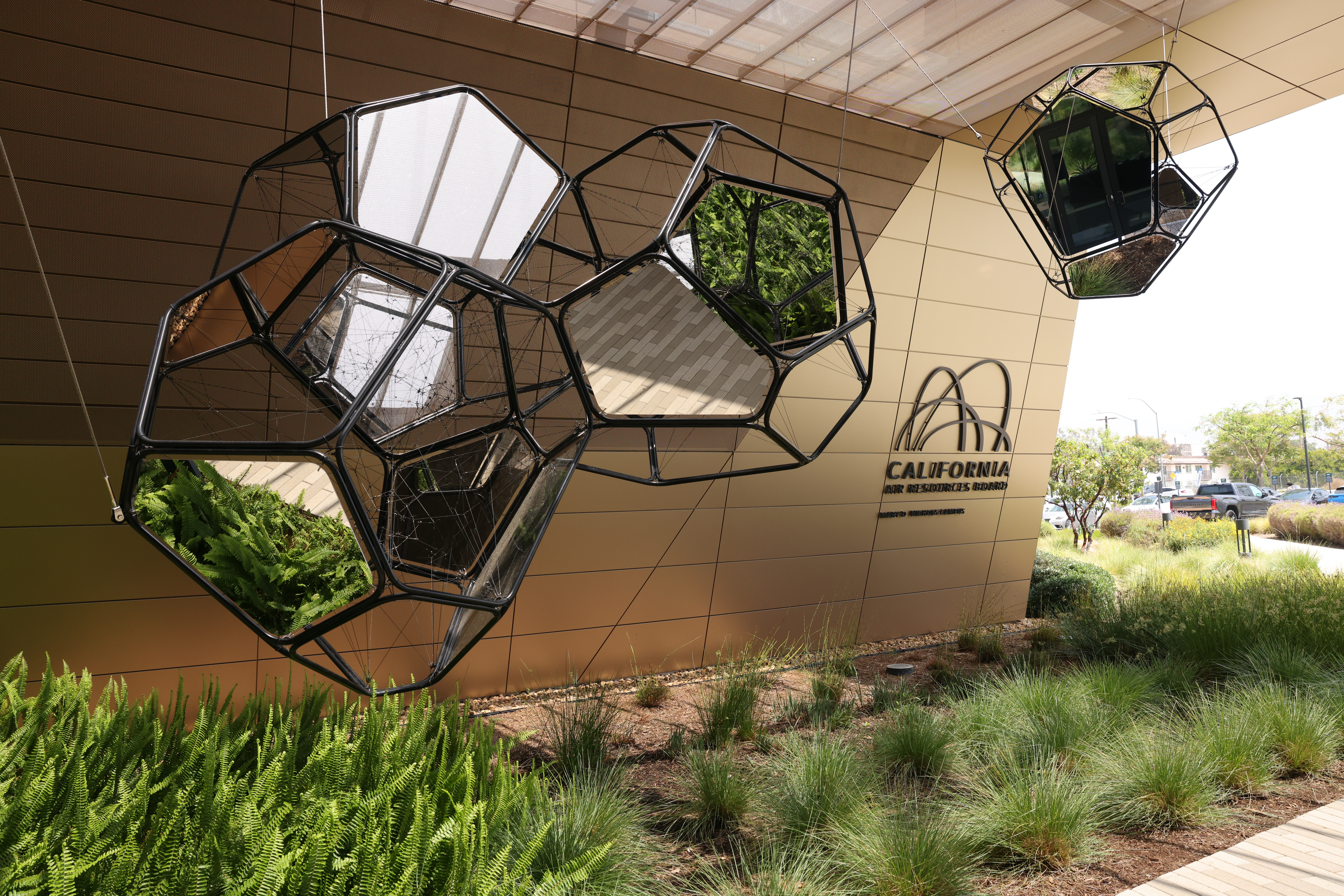 Outdoor view of the California Air Resources Board (CARB) Southern California headquarters, featuring the hanging art installation “Carbon Net,†with geometric wireframe structures and greenery integrated into the design.